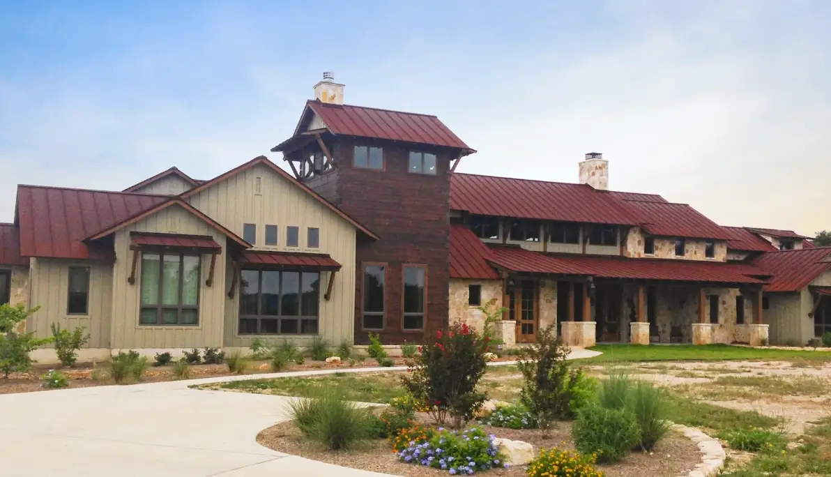 A large Hill Country style luxury home with a rust red metal roof, stone exterior, and native Texas landscaping