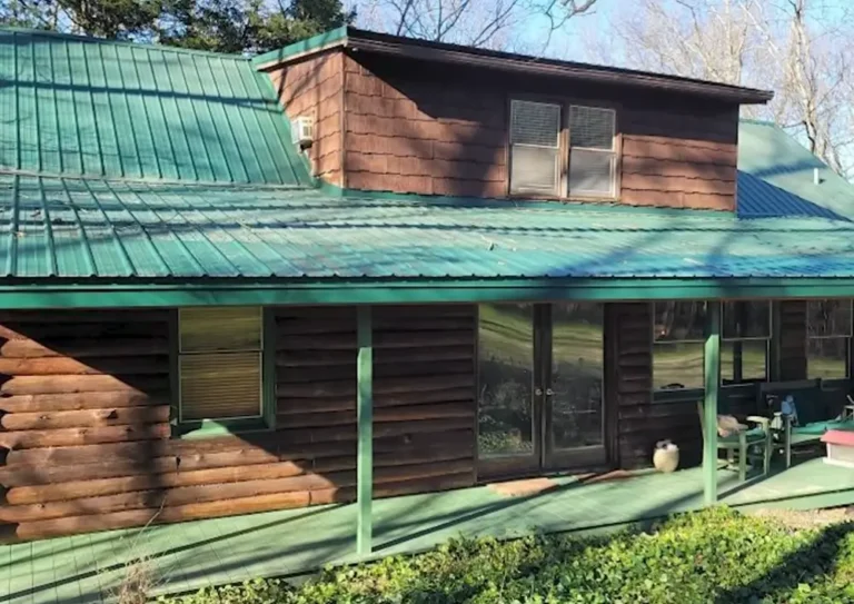 A log cabin style home with a green standing seam metal roof and a covered wraparound porch