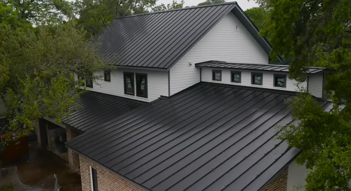 Aerial view of a home with a dark gray metal roof, showing multiple rooflines and a row of dormer windows, surrounded by mature trees