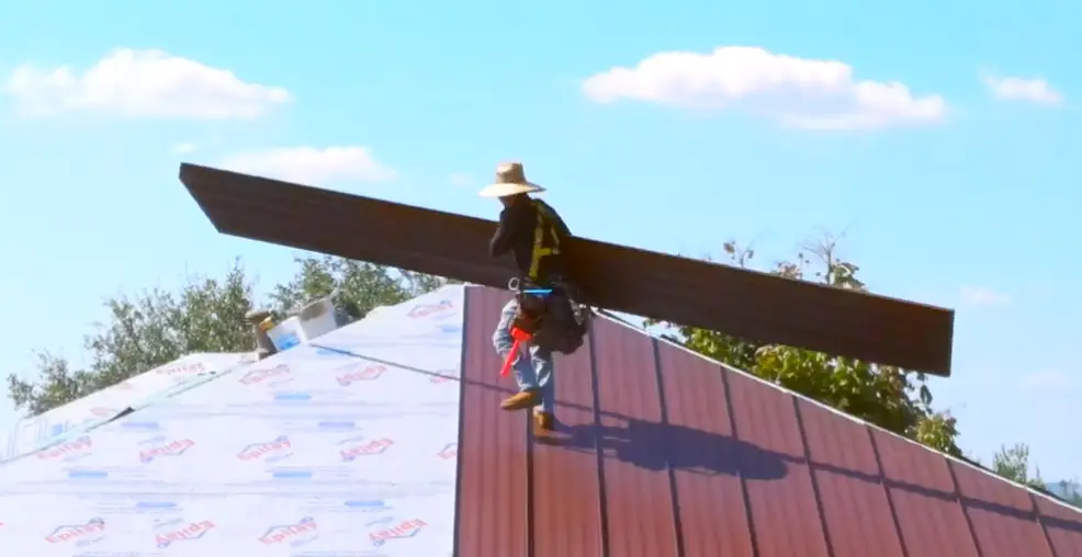 Roofer carrying a metal roofing panel up a partially installed standing seam roof with synthetic underlayment visible