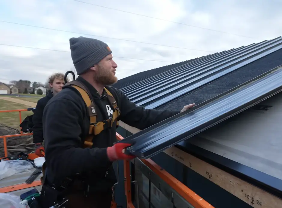 Two roofers wearing safety harnesses lift a dark standing seam metal panel onto a residential roof from a construction lift