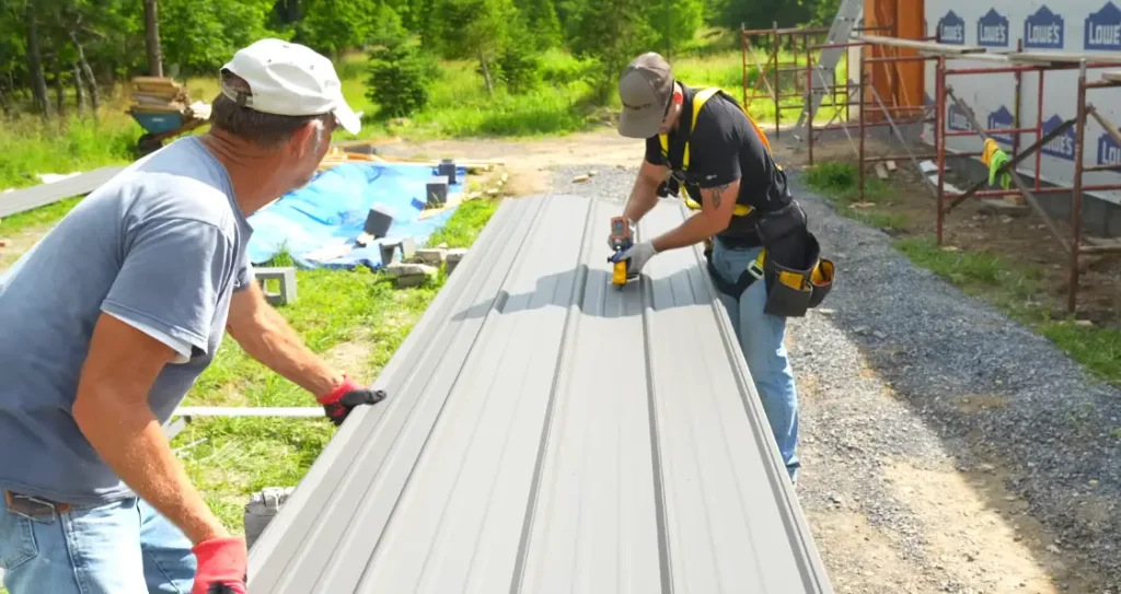 Two roofing team members holding and fastening a gray metal panel outdoors near a construction site