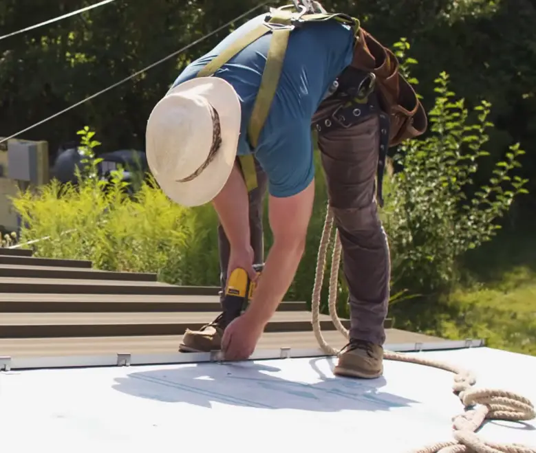 Roofing contractor wearing a safety harness uses a drill to fasten a metal standing seam panel to a roof