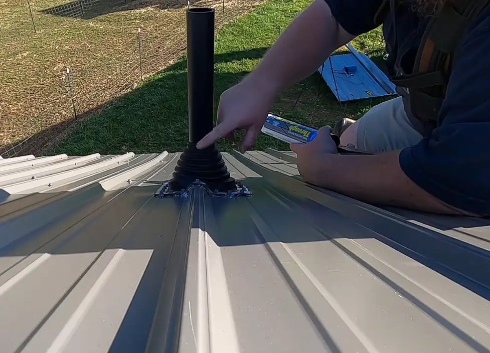 Roofer applying sealant around a black pipe boot flashing on a gray corrugated metal roof