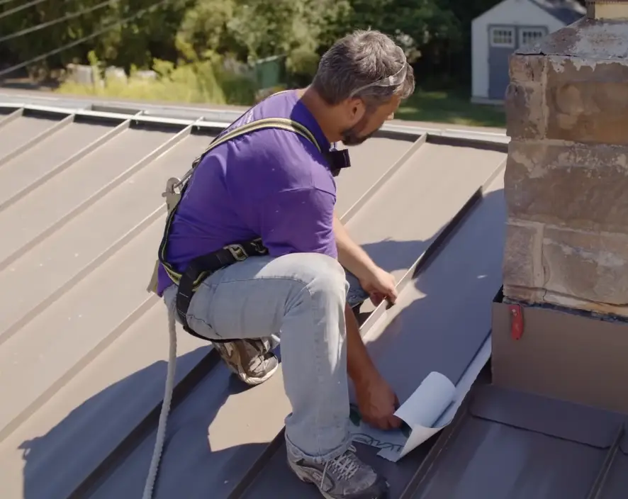 Roofer wearing a safety harness installs flashing near a chimney on a standing seam metal roof