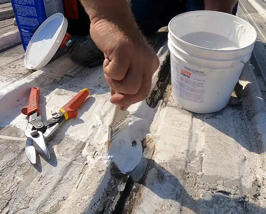 Roofer brushing white sealant compound over a seam on a metal roof, with tin snips and a sealant bucket nearby