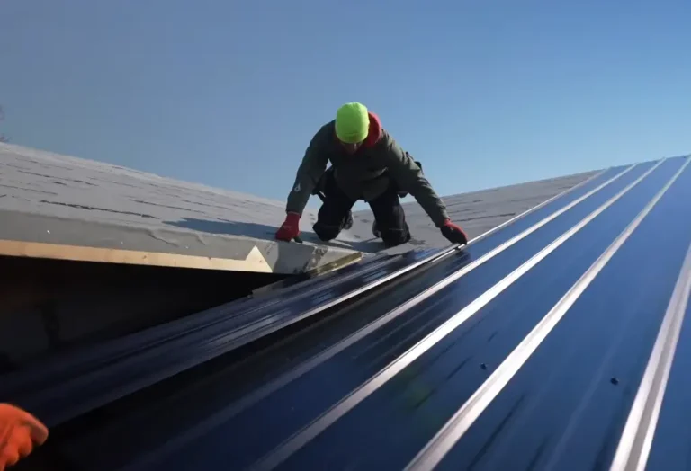 A roofer kneeling on a metal roof secures a panel near the ridge