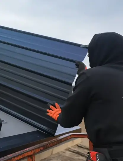 A roofer in cold weather gear installs a dark standing seam metal panel on a roof
