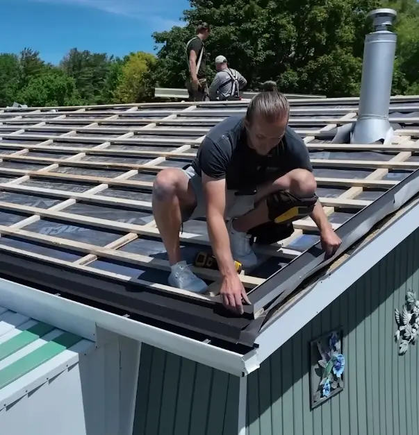 A roofing crew installs dark roof panels over battens and framing on a home, with a metal chimney vent and mature trees in the background