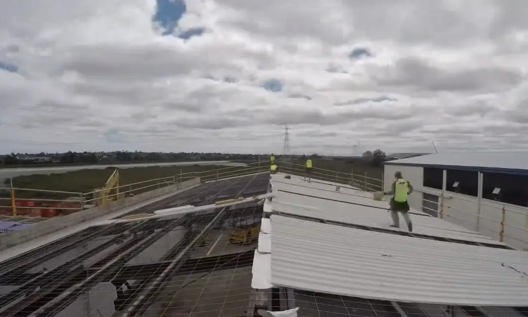 Workers in high visibility vests install corrugated metal roofing panels on a large commercial building under an overcast sky