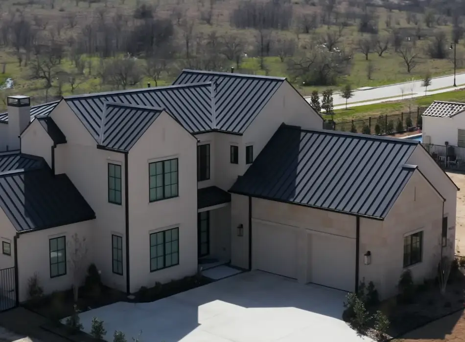 A large Hill Country style home with a dark standing seam metal roof and stone accents, seen from above with a neighboring pool visible