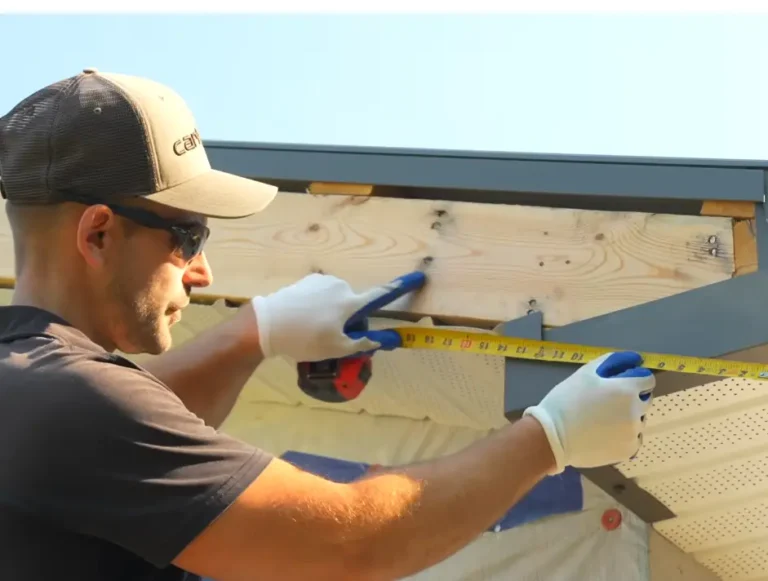 A roofer wearing safety glasses and gloves measures roofline dimensions with a tape measure during fascia and soffit installation
