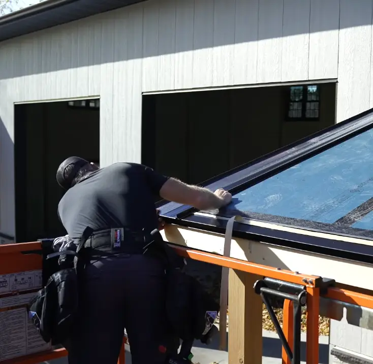 A roofer installs a blue-toned standing seam metal panel on a peaked roof, secured with a lift beside a light gray sided building
