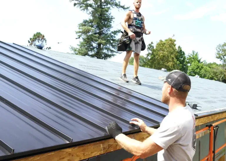 Two roofers install dark standing seam metal panels on a residential roof, one securing the panel edge, one walking the ridge