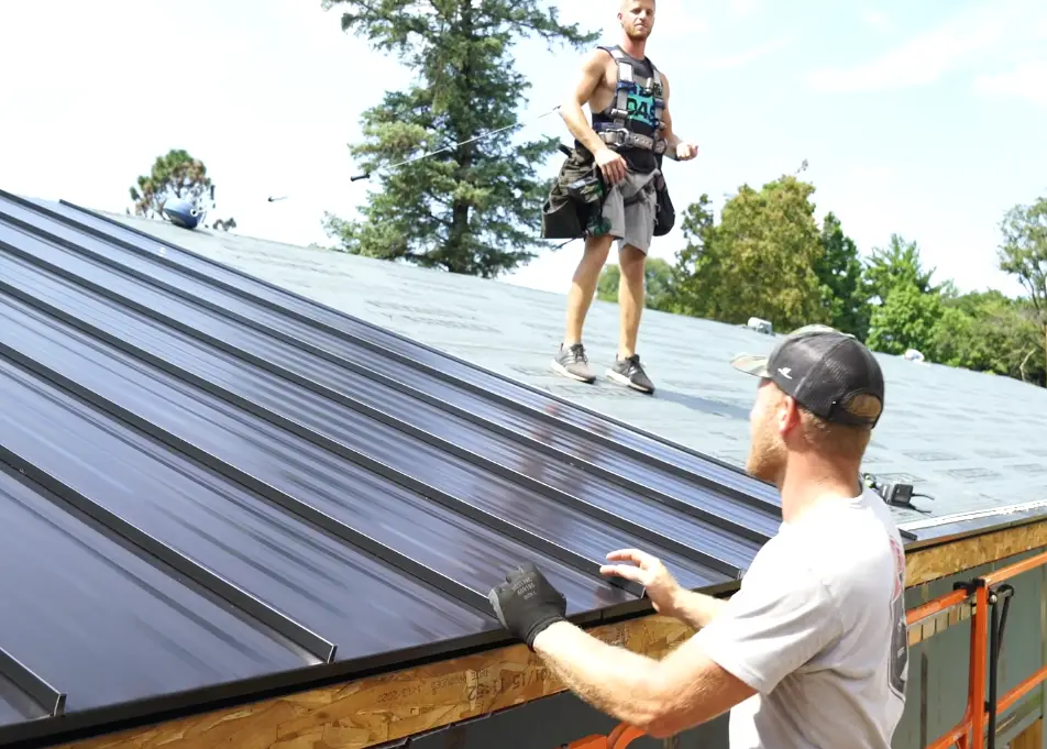Two roofers install dark standing seam metal panels on a residential roof, one securing the panel edge, one walking the ridge