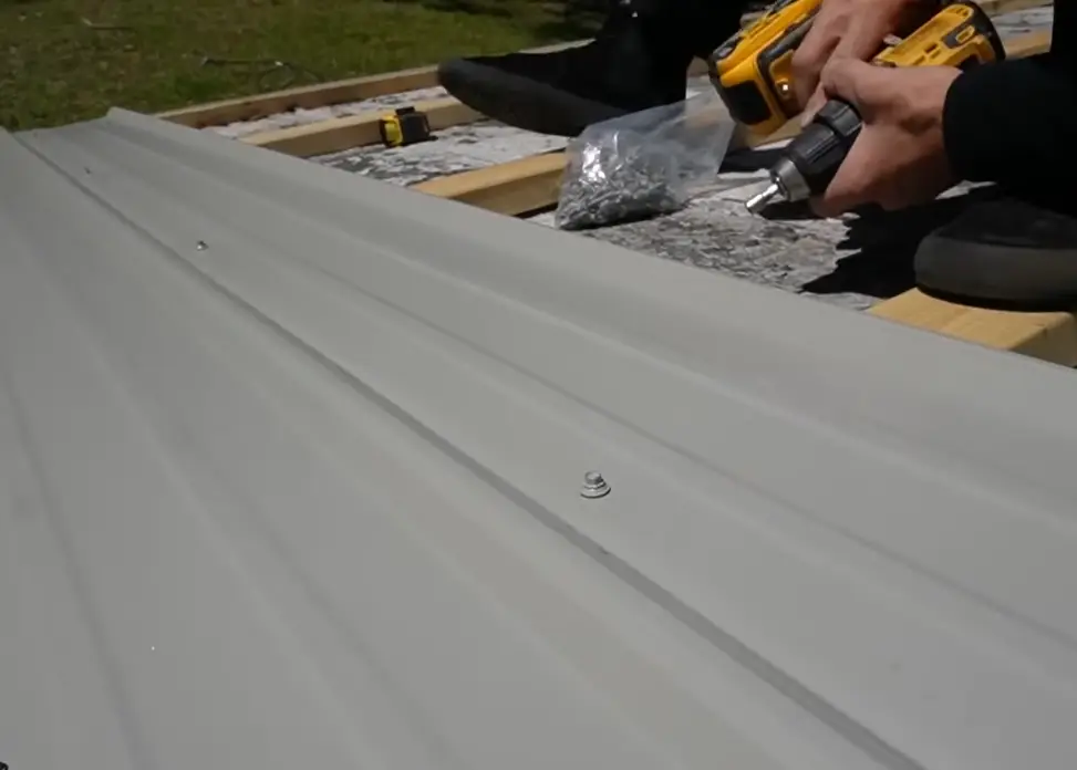 Close-up of a light gray metal roof panel being fastened with a drill, showing the exact fastener spacing that affects labor and materials cost