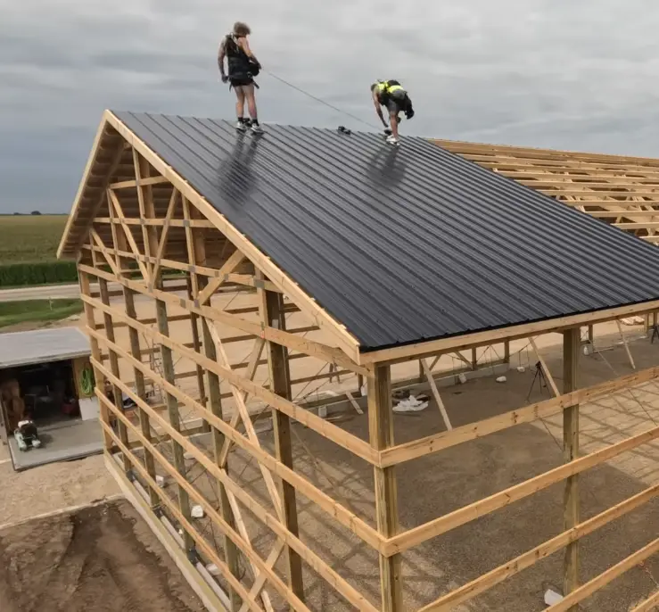 Two roofers install black corrugated metal panels on an open-frame pole barn structure surrounded by farmland