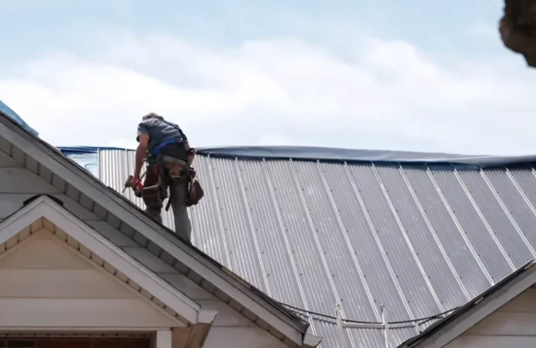 A roofer in a safety harness installs light gray metal panels on a peaked roof with light siding below