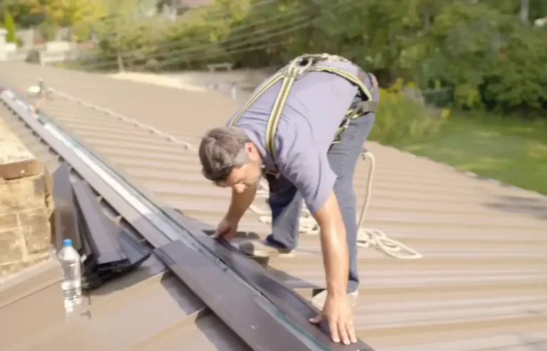 A roofer wearing a safety harness installs a standing seam metal panel edge trim near a chimney on a tan metal roof