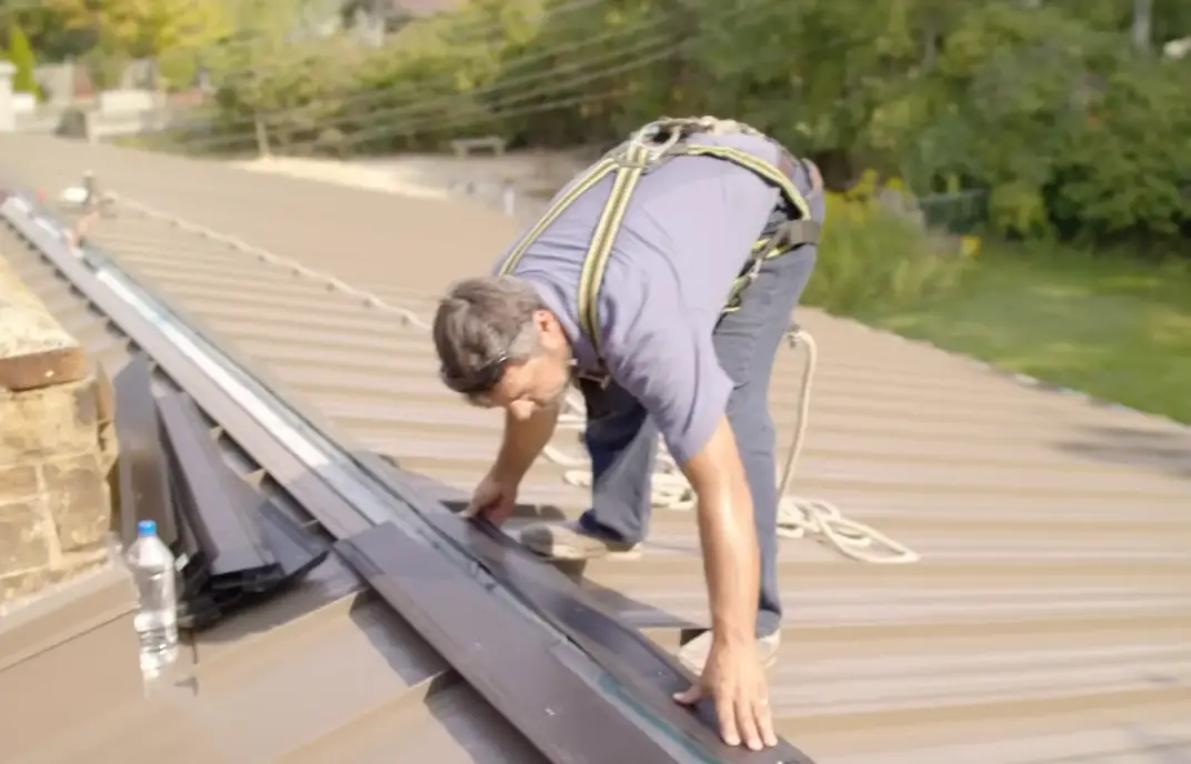 A roofer wearing a safety harness installs a standing seam metal panel edge trim near a chimney on a tan metal roof