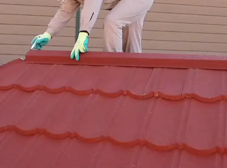 A worker's gloved hands install a red stone coated steel ridge cap on a tile profile metal roof